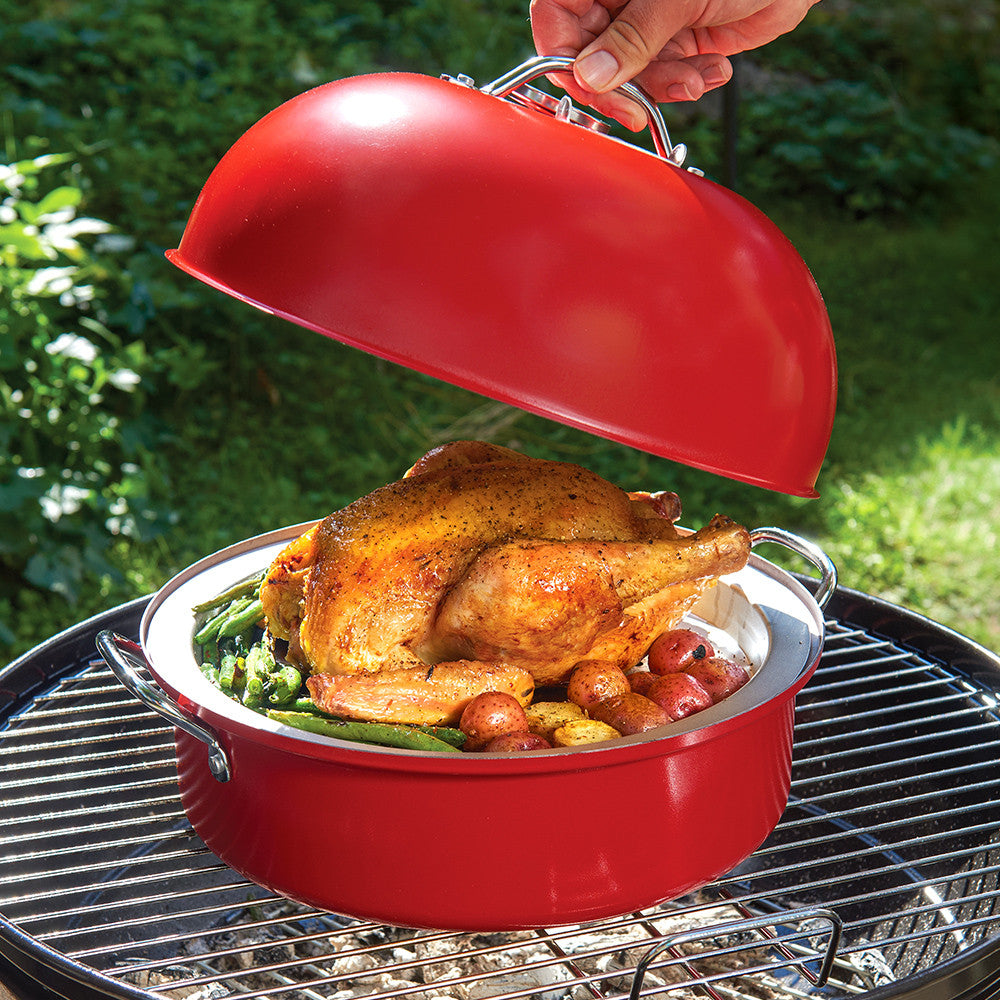 Red kettle smoker with a roasted chicken and vegetables on a grill outdoors.
