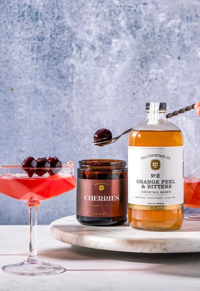 Cocktail ingredients including a bottle of 'No. 2 Orange Peel & Bitters', a jar of cherries, and a cocktail glass with cherries on a marble surface.