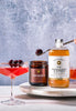 Cocktail ingredients including a bottle of 'No. 2 Orange Peel & Bitters', a jar of cherries, and a cocktail glass with cherries on a marble surface.