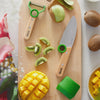 Wooden cutting board with sliced mango, kiwi, and Opinel kids kitchen tools on a light background.